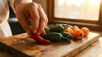Hand selecting fresh red chili pepper from wooden cutting board with green peppers and cherry tomatoes in warm kitchen lighting for cooking preparation.