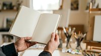 Hands holding open blank book in artist studio with paintbrushes and supplies, creative workspace for writing, journaling, or documenting artistic projects.