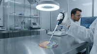 Male scientist in white lab coat conducting research with pipette in modern laboratory with petri dish containing colorful bacterial cultures.