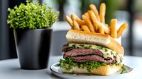 Juicy medium-rare roast beef sandwich on brioche bun topped with fresh herbs and served with golden french fries, decorative plant in background on restaurant table.