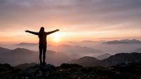 Woman hiker with arms outstretched celebrating summit achievement at sunrise over layered mountain ranges with golden sky and misty valleys below.