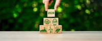 Hand placing wooden cube with 2026 text on stack of eco-friendly blocks featuring renewable energy and sustainability icons on green bokeh background.