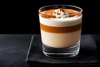 Layered coffee drink with cream foam and cocoa powder in glass on black background, moody lighting highlights distinct caramel, milk and espresso layers.