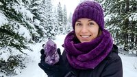 Smiling young Caucasian woman in purple winter hat and scarf holds amethyst crystal outdoors in snowy forest, spiritual wellness concept.
