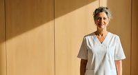 Mature woman with gray hair and medium skin tone wearing white medical scrubs standing against wooden wall with natural sunlight for healthcare professional portraits.