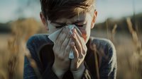 Young boy with tissue blowing nose in field of dry grass during sunset, showing seasonal allergies or cold symptoms in outdoor rural setting.