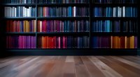 Library bookshelf with colorful book spines arranged by hue from red to blue creating rainbow gradient effect against dark background on wooden floor.