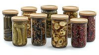 Glass jars with preserved vegetables, mushrooms and berries arranged in row showing variety of homemade canned foods with metal lids on white background.