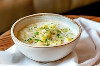 Creamy potato soup garnished with fresh chives and melted butter in white ceramic bowl, served on wooden table with linen napkin. Homestyle comfort food.