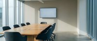 Modern corporate conference room with wooden table, black chairs and wall-mounted screen for business meetings and presentations in office building.