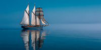 Vintage sailing ship with white sails gliding across calm blue waters, creating perfect mirror reflection against serene horizon at dusk.