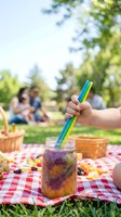 Child's hand holding colorful straw in beverage jar during outdoor picnic gathering with family and friends in sunny park setting.
