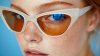 Close up portrait of young woman with freckles wearing trendy cat eye sunglasses with orange lenses against blue background. Fashion and beauty concept.