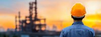 Industrial engineer in safety helmet observing oil refinery plant at sunset, panoramic view with silhouette of petrochemical facility against orange sky.