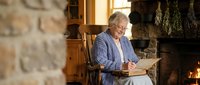 Elderly Caucasian woman in blue striped pajamas sitting by stone fireplace reading book in cozy rustic interior with warm lighting for relaxation content.