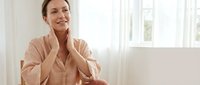 Young woman in beige robe touching face with gentle smile in bright bathroom, skincare routine, morning self-care ritual, wellness and beauty concept.