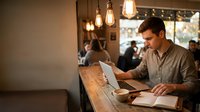 Young male professional of mixed ethnicity working at wooden counter with laptop and documents in warm-lit cafe or office space with pendant lighting and blurred background patrons.