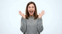 Excited young Caucasian woman with brown hair raising hands up in surprise gesture wearing gray sweater on white background isolated for design.