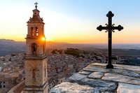 Ancient stone church bell tower and ornate cross silhouetted against dramatic sunset sky over Mediterranean hillside town, creating spiritual atmosphere.