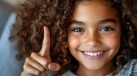 Young African American girl with curly hair and bright smile showing thumbs up gesture against blurred background. Positive expression and natural lighting.