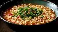 Spicy Asian noodle soup garnished with fresh chopped green onions and chili flakes in dark ceramic bowl, dramatic lighting highlights steam and texture.