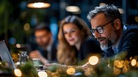 Mature businessman with glasses working on laptop during evening meeting with colleagues in restaurant atmosphere with bokeh lights.
