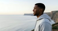 Young man of African descent wearing light blue hoodie gazing at coastal cliffs and calm ocean waters during golden hour. Suitable for lifestyle, travel, wellness, and inspirational content.