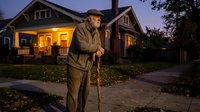 Senior Caucasian man with walking cane standing on sidewalk in front of illuminated suburban home during twilight evening hours for healthcare concepts.
