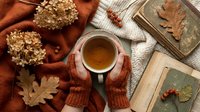 Cozy autumn flat lay with female hands in knit gloves holding a cup of hot tea, dry leaves, books, and hydrangea flowers on warm textile.