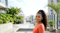 Young woman with curly dark hair and medium skin tone wearing coral top on urban rooftop terrace with potted plants and city skyline background for lifestyle and portrait use.