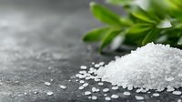 Heap of white sea salt crystals scattered on dark stone surface with fresh green herbs, macro shot. Food photography for culinary and healthy lifestyle concepts.