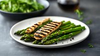 Grilled salmon fillet served with fresh green asparagus spears on white plate against dark background, garnished with black pepper and herbs.