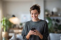 Young Caucasian woman in cozy gray sweater smiling while texting on smartphone in modern kitchen interior with warm lighting and plants.