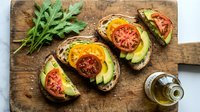 Toasted bread slices topped with ripe avocado, fresh tomato slices and melted cheese on rustic wooden cutting board with arugula and olive oil for healthy breakfast or lunch.