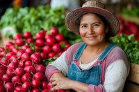 Smiling middle-aged Hispanic woman vendor in straw hat and overalls selling fresh radishes and vegetables at local farmers market.
