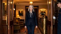 Professional blonde businesswoman in navy suit walking through luxury hotel lobby with elegant wood paneling and warm lighting atmosphere.