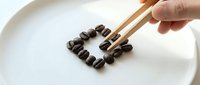 Caucasian hand holding chopsticks arranging roasted coffee beans into a heart shape on a white ceramic plate, minimalist style.