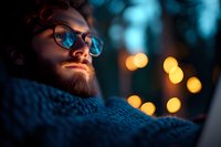 Bearded man in glasses illuminated by warm bokeh lights against dark blue evening background, wearing knit scarf, contemplative winter portrait.