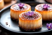 Mini caramel flan desserts garnished with purple edible flowers on dark plate, close up view showing creamy texture and caramelized sugar topping with artistic presentation.