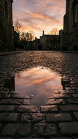 Historic stone courtyard with puddle reflecting golden sunset sky, bare trees and medieval architecture, ideal for travel and heritage website backgrounds.