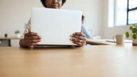 Man with dark skin tone holding blank white paper at wooden desk in bright office space with plants and window background.