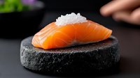 Fresh salmon sashimi slice topped with sea salt crystals on black stone plate against dark background, dramatic lighting highlights texture and color.