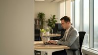 Professional businessman working on laptop in modern office space with natural lighting and plants creating productive workspace atmosphere.