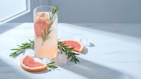 Refreshing grapefruit rosemary cocktail with ice in tall glass on white marble surface. Fresh citrus slices and herb garnish create elegant drink presentation.