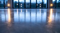 Atmospheric evening interior with polished concrete floor reflecting warm lights from large windows, creating symmetrical glowing pattern in empty modern space.