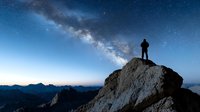 Lone hiker silhouette standing on mountain peak under spectacular Milky Way galaxy stretching across starry night sky with mountain ranges below.