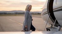 Confident mature blonde businesswoman in gray coat boarding private jet at sunset on airport tarmac for luxury corporate travel and executive lifestyle.