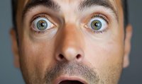 Close up portrait of surprised male face with wide open blue eyes and mouth expressing shock and amazement against gray background.