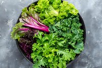 Fresh mixed salad greens with purple radicchio, curly lettuce, and kale varieties arranged in black bowl on gray concrete background, top view.
