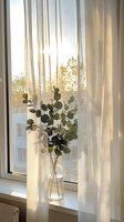 Eucalyptus branches in glass vase on windowsill with soft morning sunlight filtering through sheer curtains creating warm home atmosphere.
